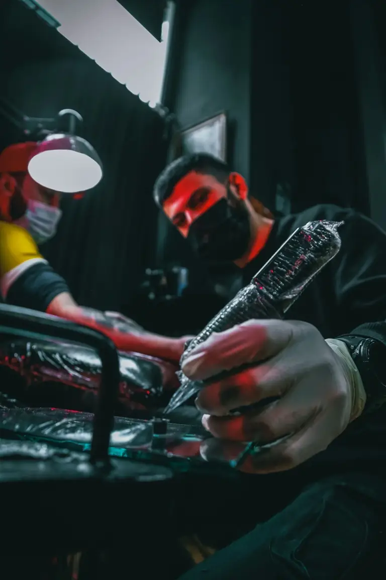 Masked artist under red light holding a wrapped tattoo pen